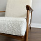 A close-up of a mid-century modern armchair with textured off-white bouclé upholstery and a warm medium-brown wooden frame, set against a neutral gray wall and dark wood floor.