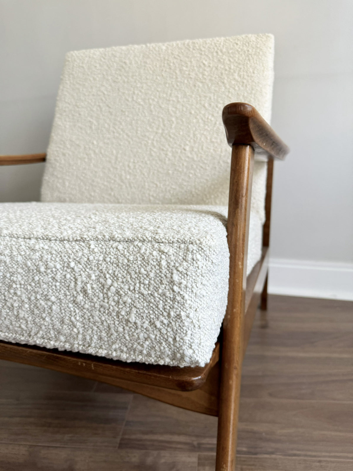 A close-up of a mid-century modern armchair with textured off-white bouclé upholstery and a warm medium-brown wooden frame, set against a neutral gray wall and dark wood floor.