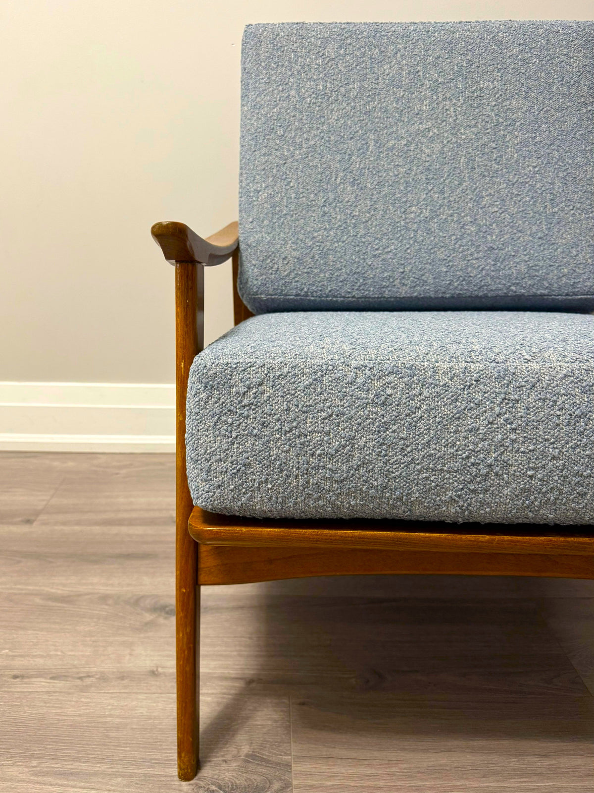 A close-up of a mid-century modern armchair with textured light blue-gray bouclé upholstery and a warm medium-brown wooden frame, set against a neutral beige wall and gray wood-look floor.