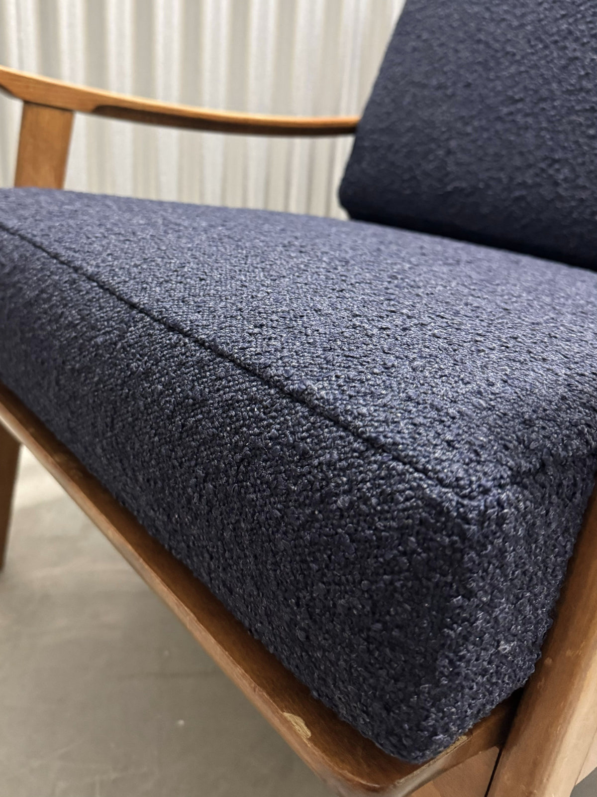 Close-up of a mid-century modern armchair with a dark navy blue textured bouclé fabric seat cushion and a light natural wood frame.