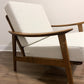 A mid-century modern accent chair with a warm brown wooden frame and textured off-white fabric cushions, shown on a wood floor against a plain wall.