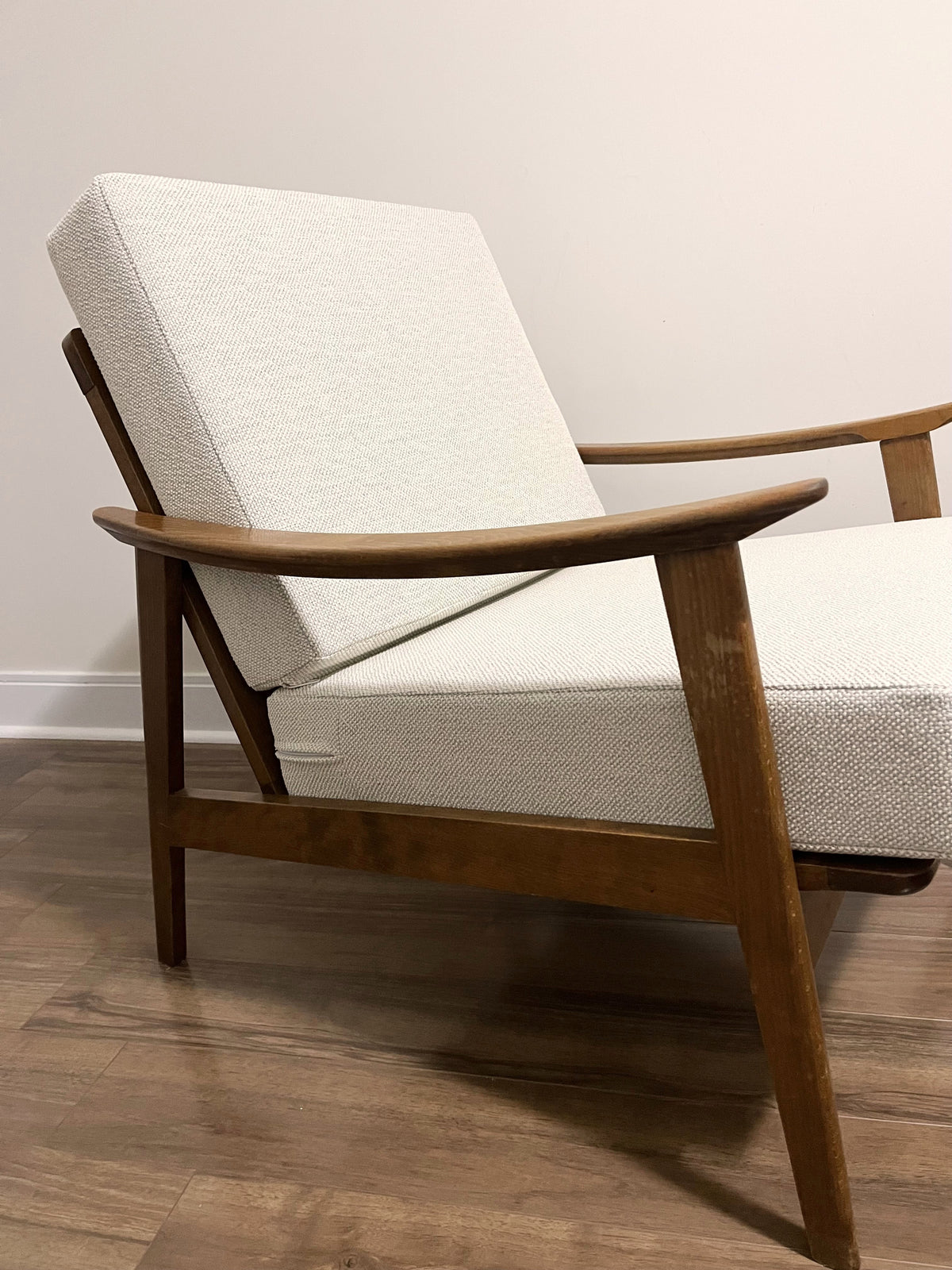 A mid-century modern accent chair with a warm brown wooden frame and textured off-white fabric cushions, shown on a wood floor against a plain wall.