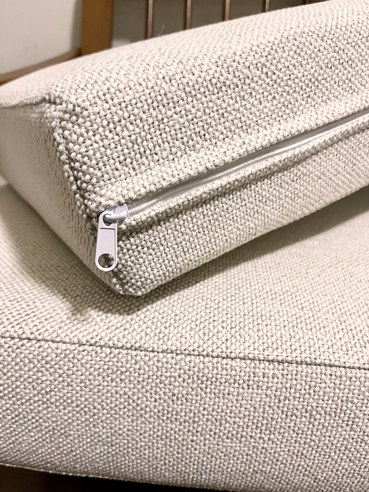Close-up of a cream woven fabric cushion with a white zipper pull, resting on a matching cushion.