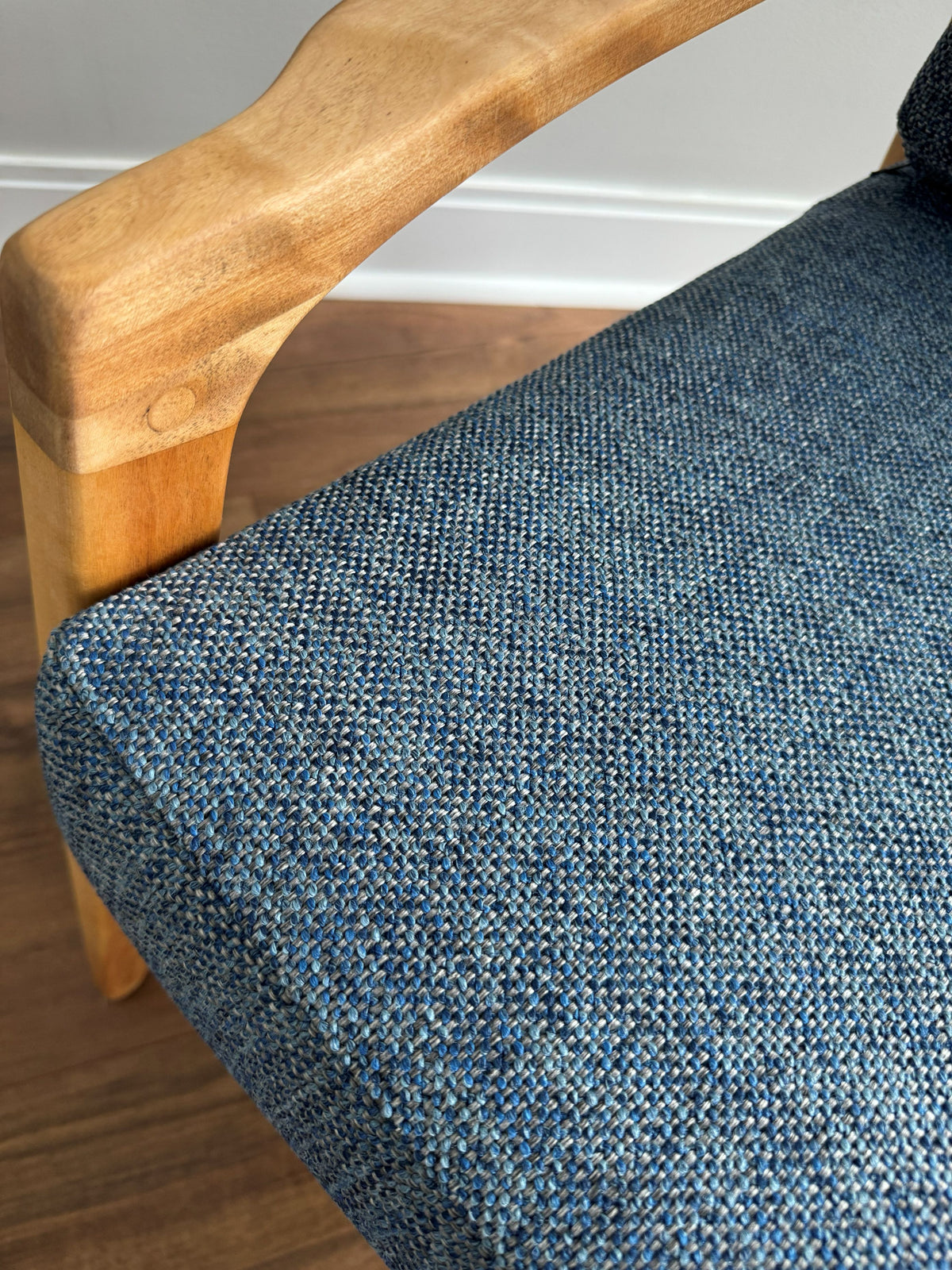 Close-up of a blue textured woven upholstery fabric on an armchair, with a light natural wood armrest and wooden floor visible.