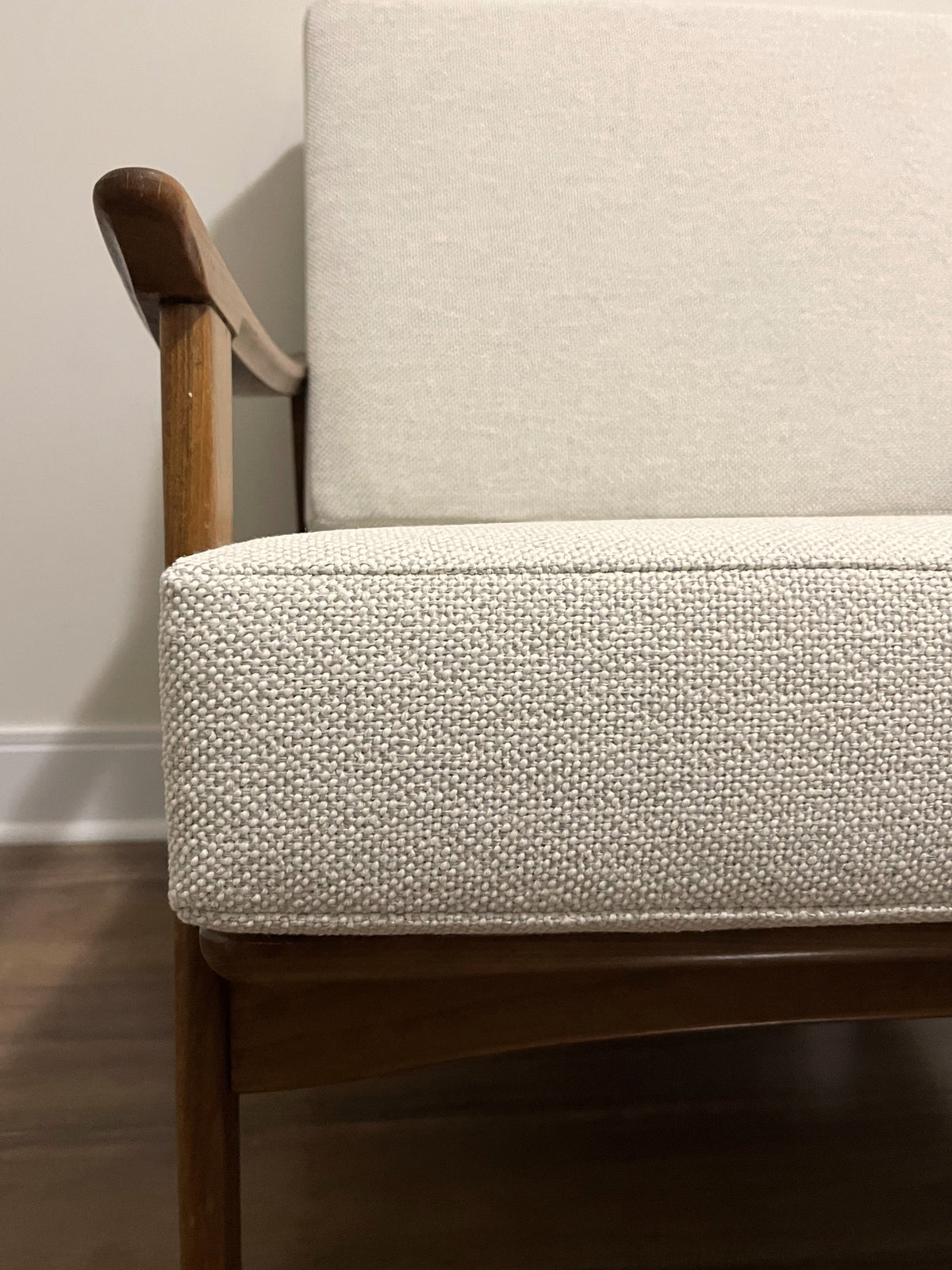 Close-up of a mid-century modern armchair with textured off-white woven upholstery and a warm medium-brown wooden frame.